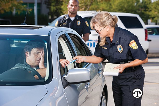 A mature female law enforcement officer stands by a vehicle she has stopped and takes the driver's license from a middle aged man while her partner stands by in the background.